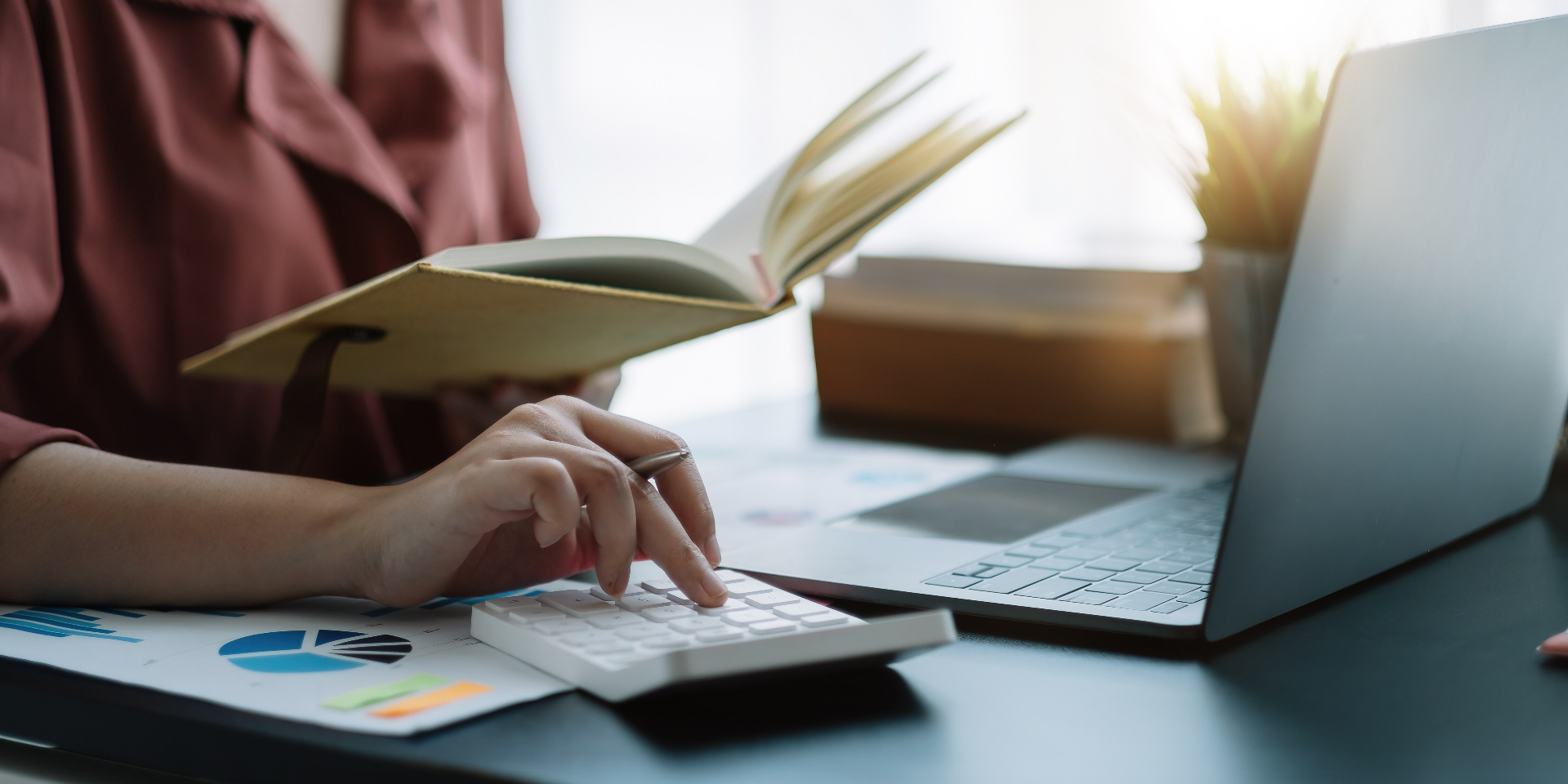 Bookkeeper reconciling monthly close with calculator, ledger, and laptop